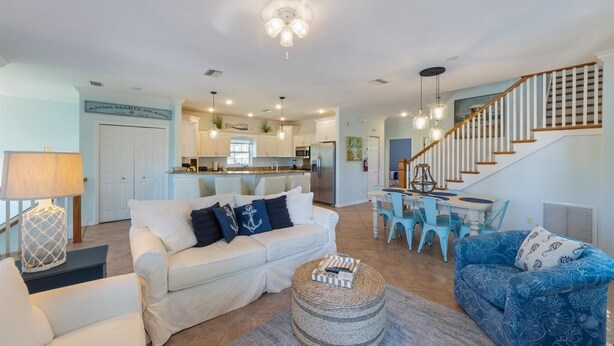 A spacious living area with a sofa, ottoman, and dining table. A kitchen with white cabinets and a staircase is visible in the background. - Blue Haven Beach House: 3BR/3BA Pool Home Steps to Beach (Holmes Beach)