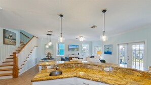 A spacious kitchen with granite countertops, white cabinetry, and a central island. There is a wooden staircase leading to another level, and large windows providing natural light. - Blue Haven Beach House: 3BR/3BA Pool Home Steps to Beach (Holmes Beach)