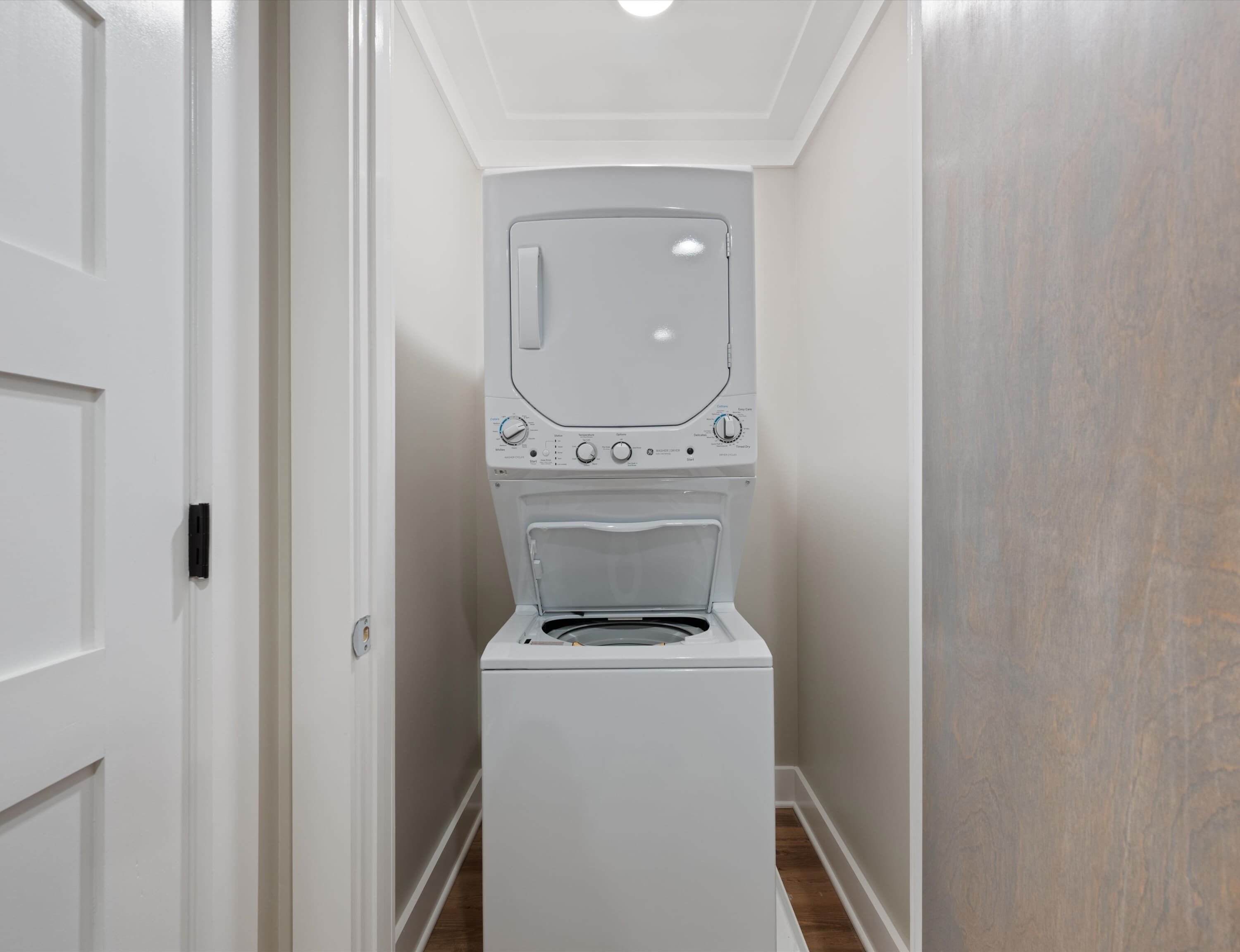 A laundry room with a stacked washer and dryer, a white door, and a wooden floor.