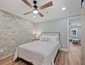 Un dormitorio con pared de ladrillo, suelo de madera, cama con colcha blanca, mesita de noche con lámpara y ventilador de techo.