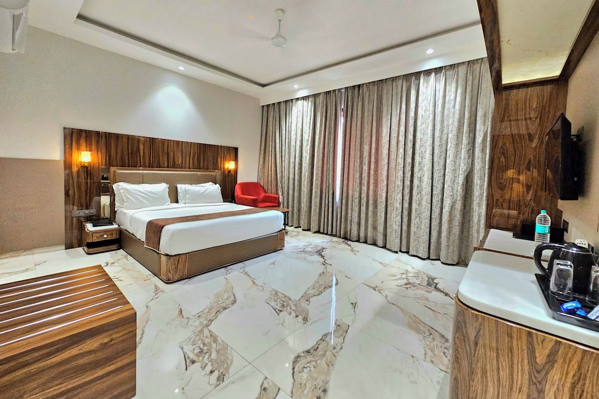 Een hotelkamer met een groot bed, een houten hoofdeinde, een rode stoel, een nachtkastje van hout, een marmeren vloer, een groot raam met gordijnen en een klein kitchenette.
