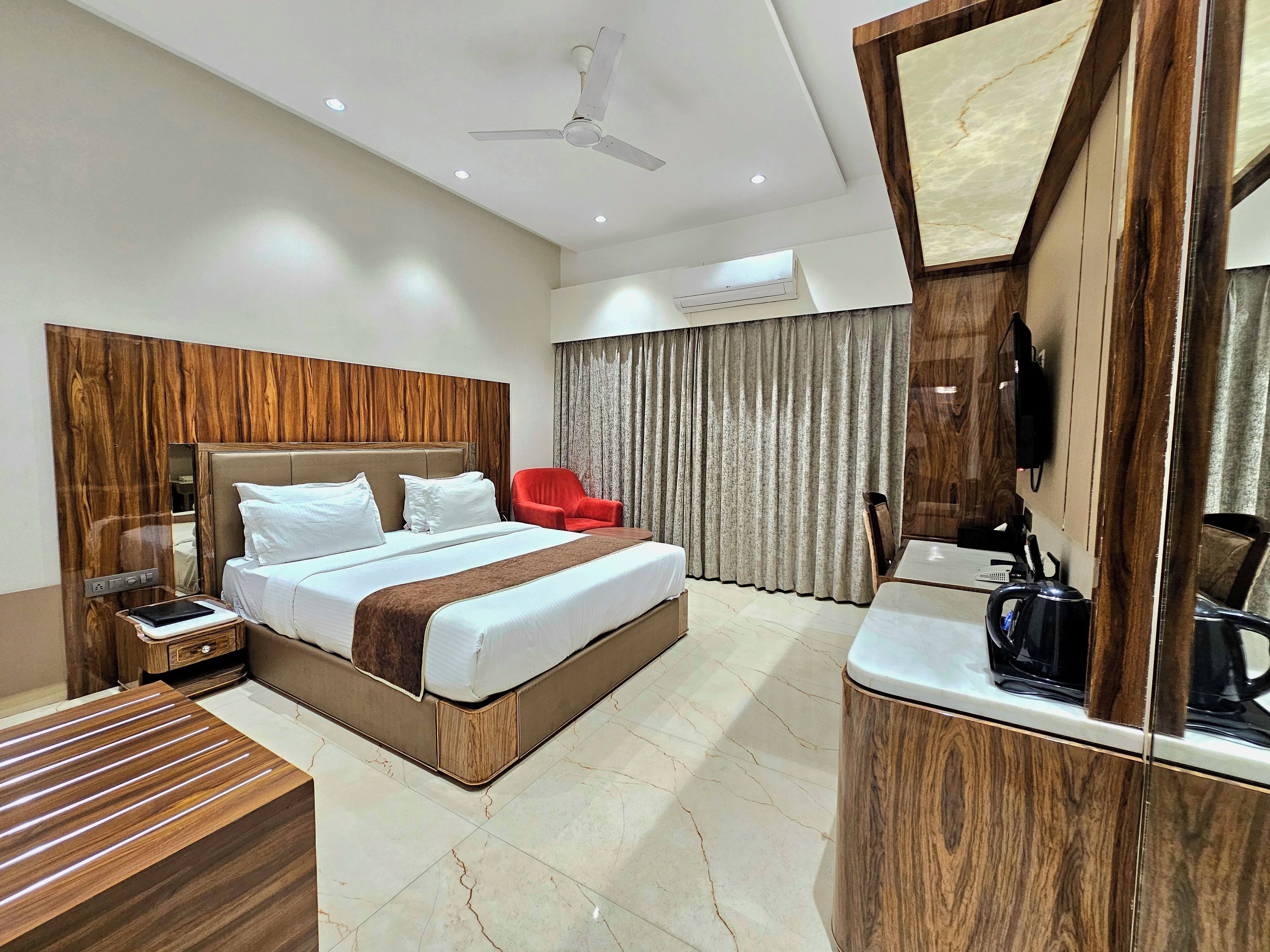 A modern hotel room with a large bed, a wooden headboard, a red armchair, a bedside table with a lamp, and a built-in TV.