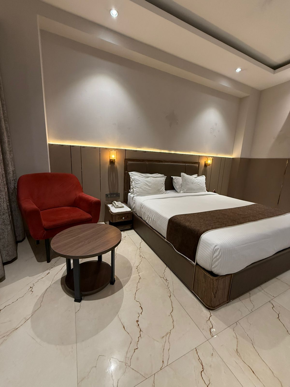 Una habitación de hotel con una cama grande, un sillón rojo, una mesita redonda y una mesita de noche con un teléfono.