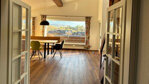An open-plan room with a wooden floor, a dining table with chairs, a hanging pendant light, and a view of buildings and trees through large windows. - Holiday Apartment 'Nini's Berge' with Panoramic View, Shared Garden and Wi-Fi (Sonthofen)