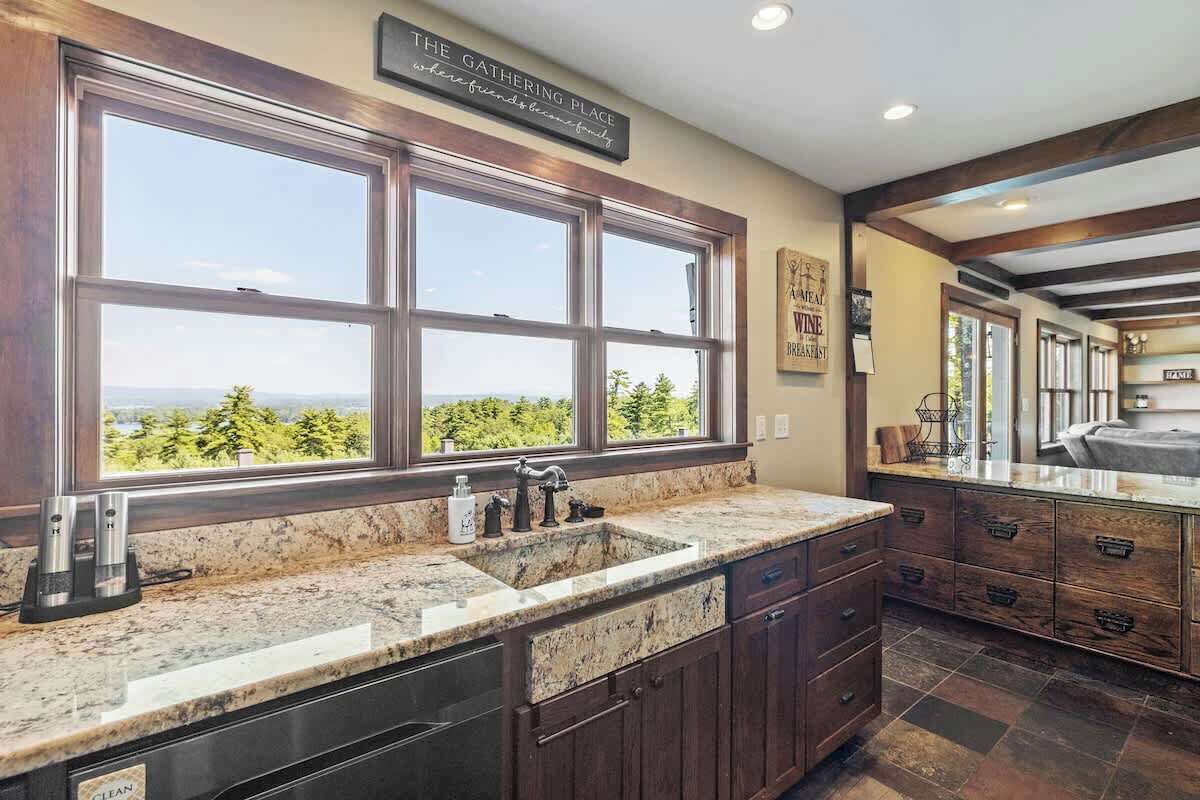 A kitchen with a large window, granite countertops, dark wooden cabinets, and a view of greenery.
