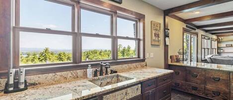 A kitchen with a large window, granite countertops, dark wooden cabinets, and a view of greenery.