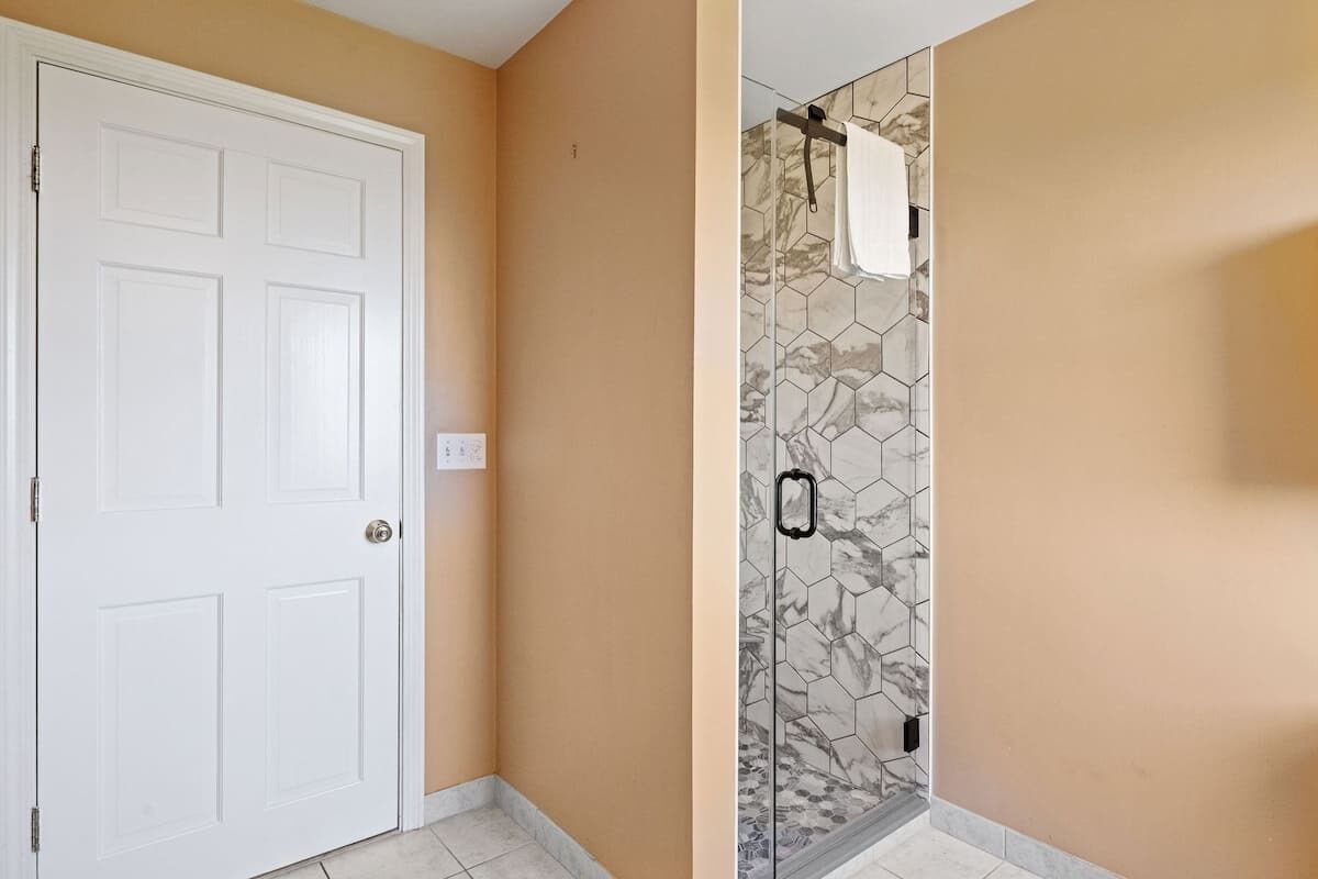 Un baño con puerta blanca, ducha con azulejos hexagonales y pared beige.