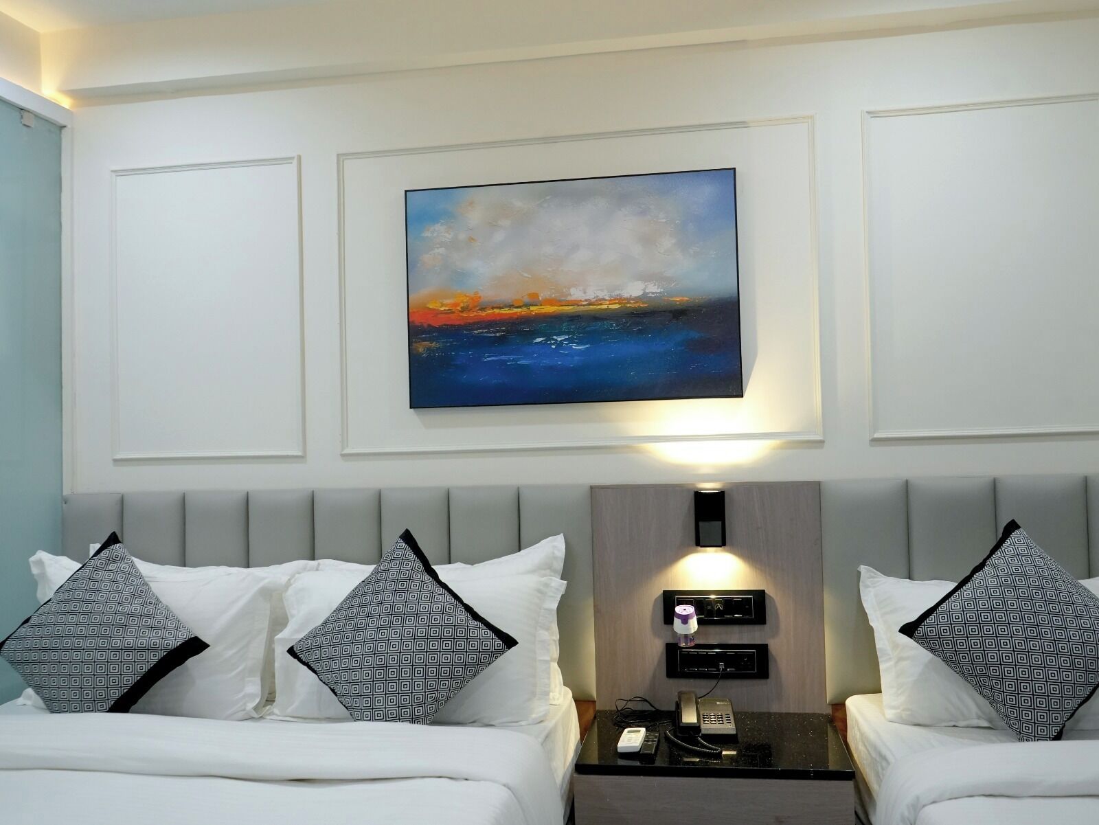 A hotel room with a bed, two bedside tables, a painting of a sunset, and a wall-mounted phone.