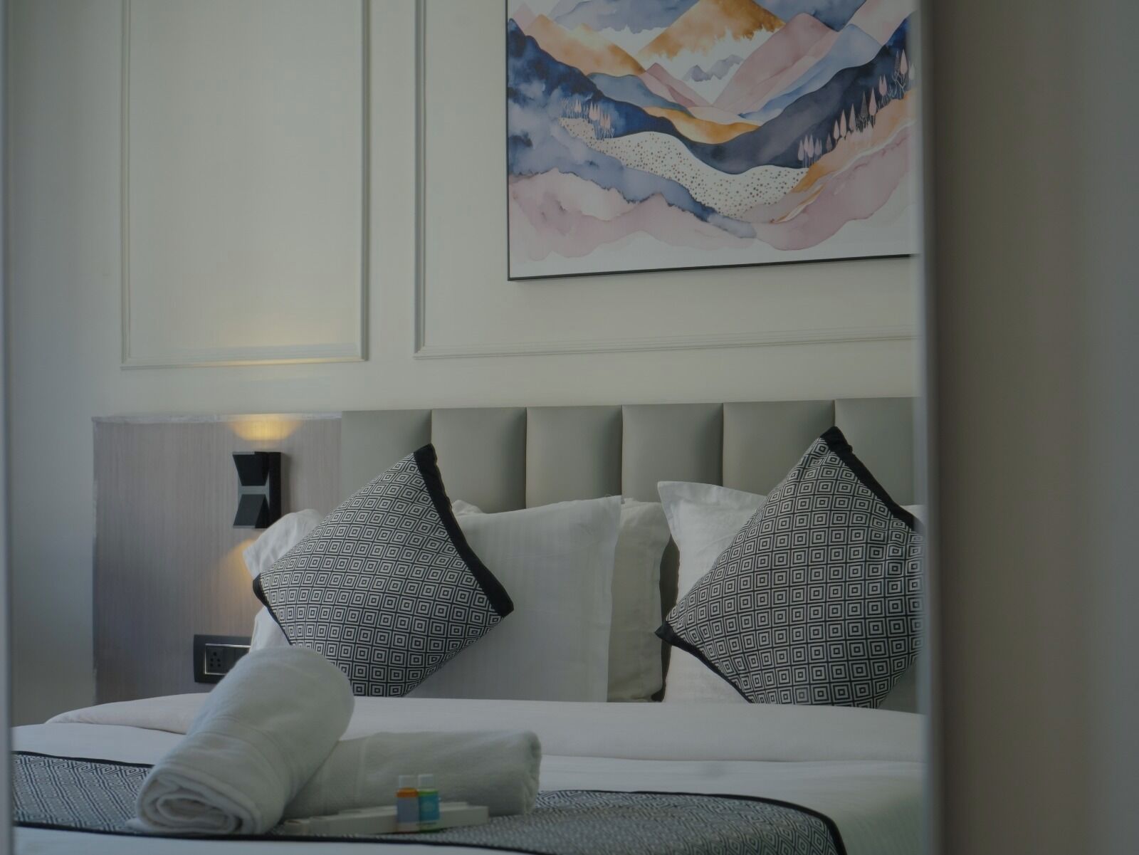 Una habitación de hotel con una cama, almohadas, una obra de arte en la pared y una mesita de noche con lámpara.