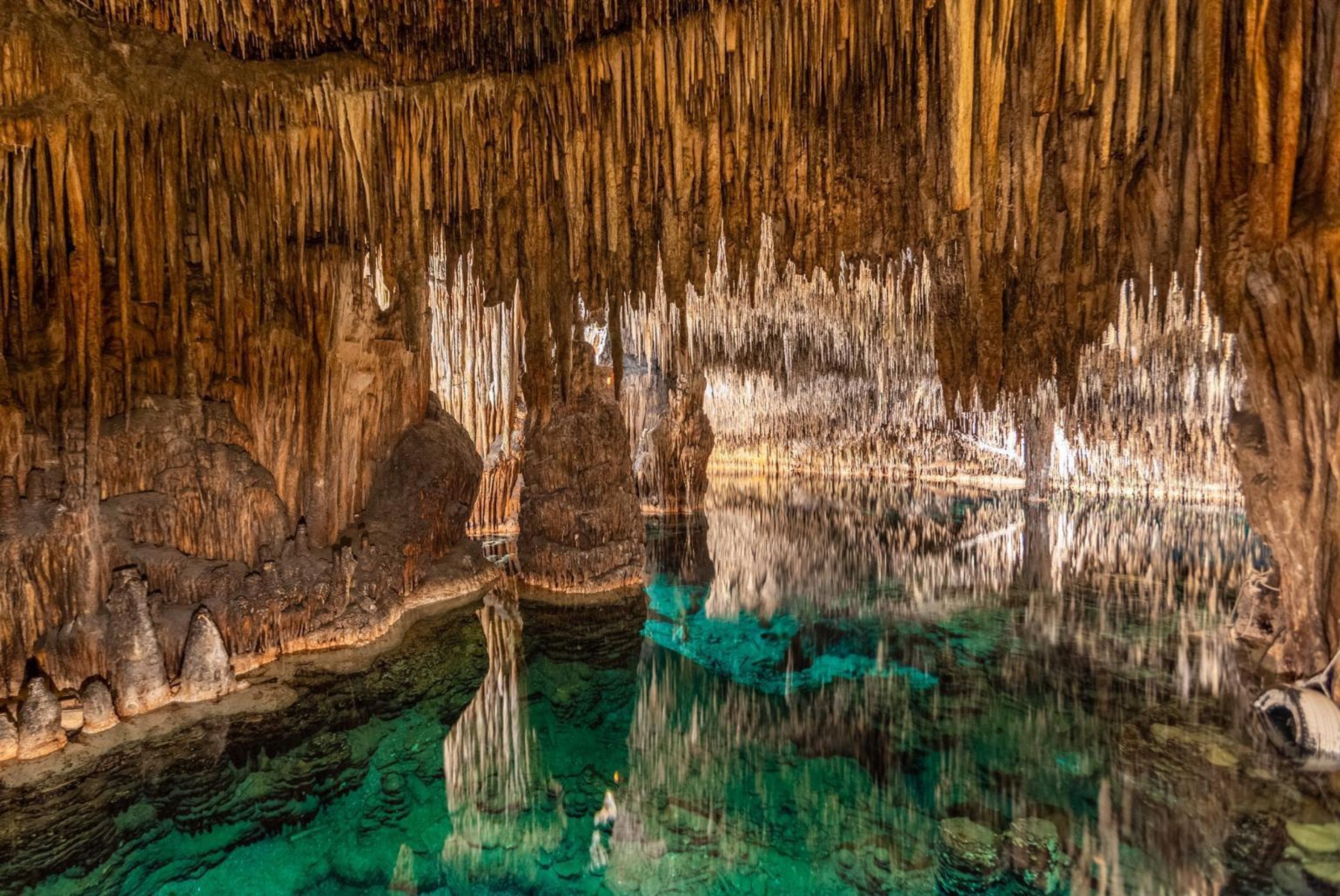 Interior de una cueva con estalactitas y estalagmitas, agua cristalina que refleja las formaciones y un suelo rocoso.