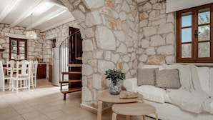 Un amplio salón con una pared de piedra, un sofá blanco, una mesa de comedor y una escalera que conduce a otra habitación.