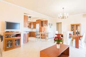 A modern kitchen with wooden cabinets, a dining area with a table and chairs, and a living room with a TV and a bench.
