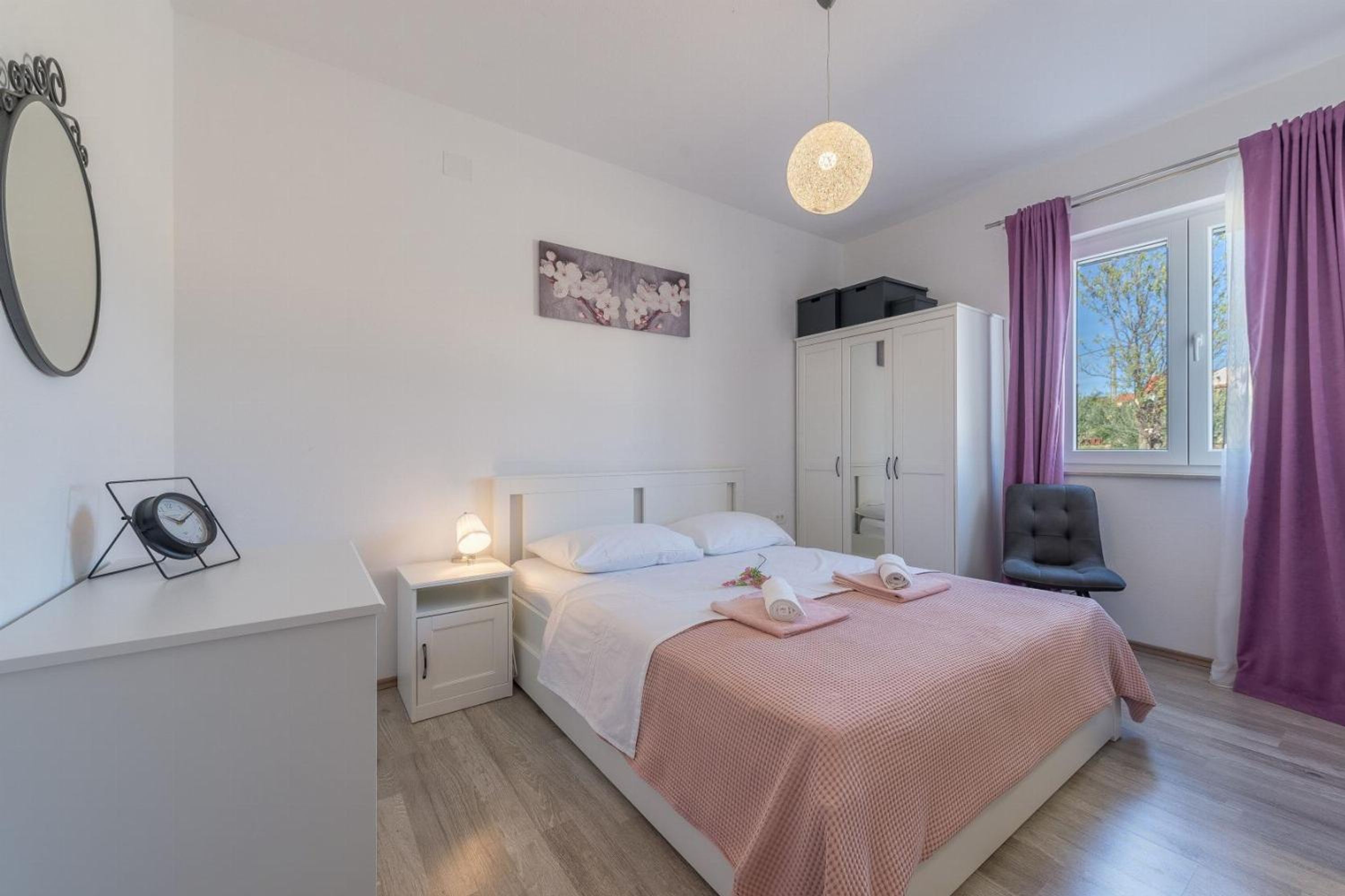 Un dormitori amb llit, taula de nit, làmpada, armari, cadira i finestra amb cortines.