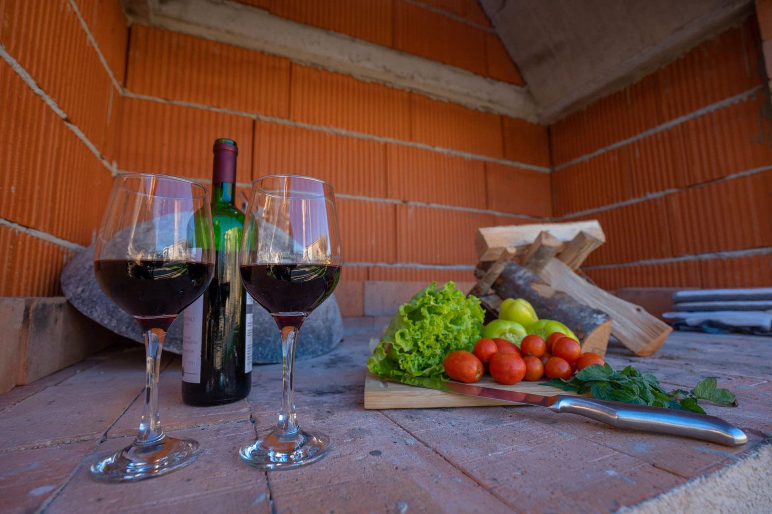 Twee wijnglazen gevuld met rode wijn, een fles, en een houten snijplank met groenten en een mes op een bakstenen oppervlak.