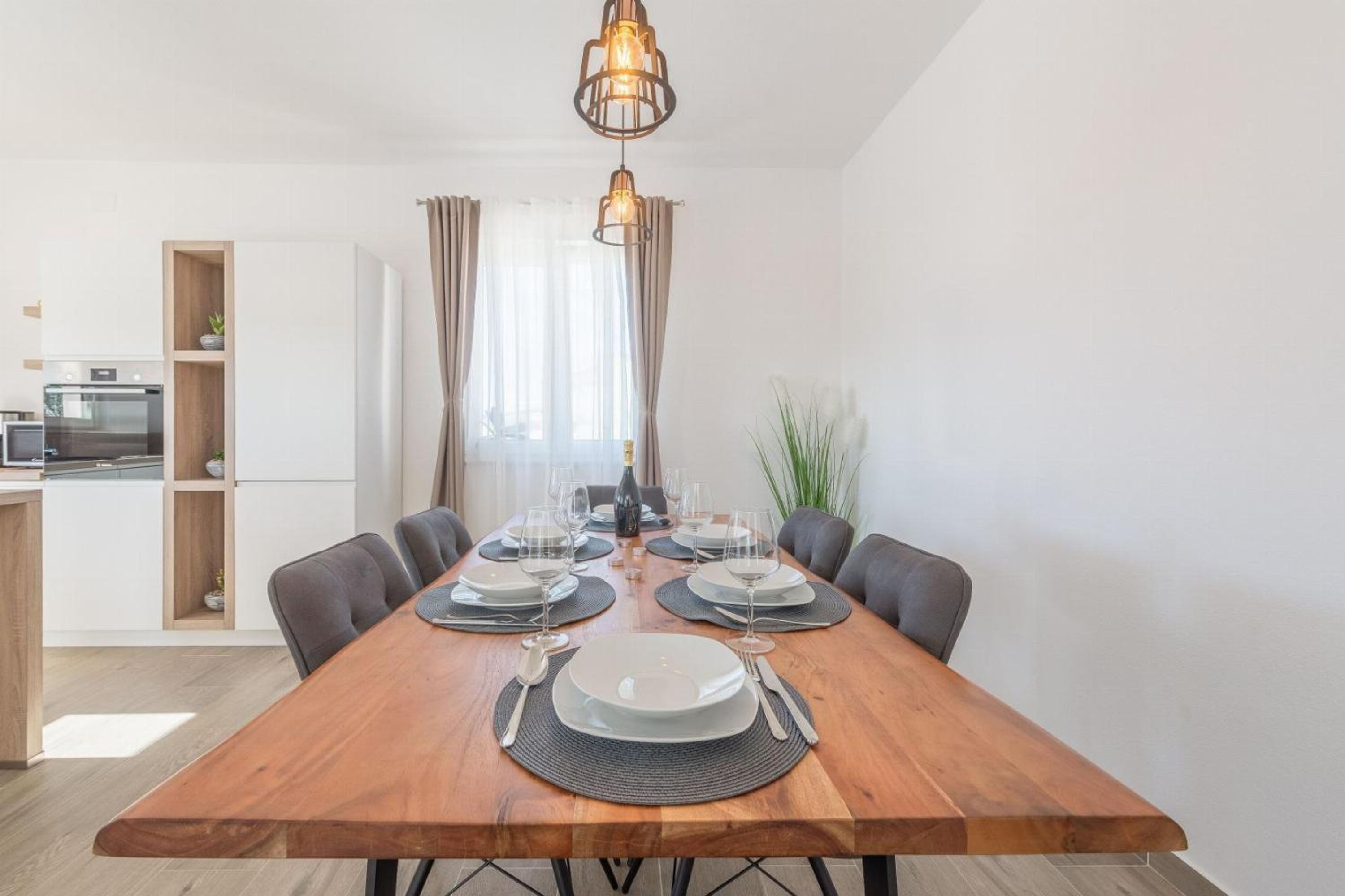 A dining area with a wooden table set for four, surrounded by chairs, with a kitchen in the background.
