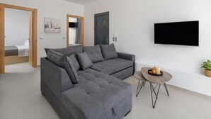 A modern living room with a grey sectional sofa, a flat-screen TV mounted on the wall, and a small round table with candles. - Salty Sun 4 in Kremasti (Rhodes)