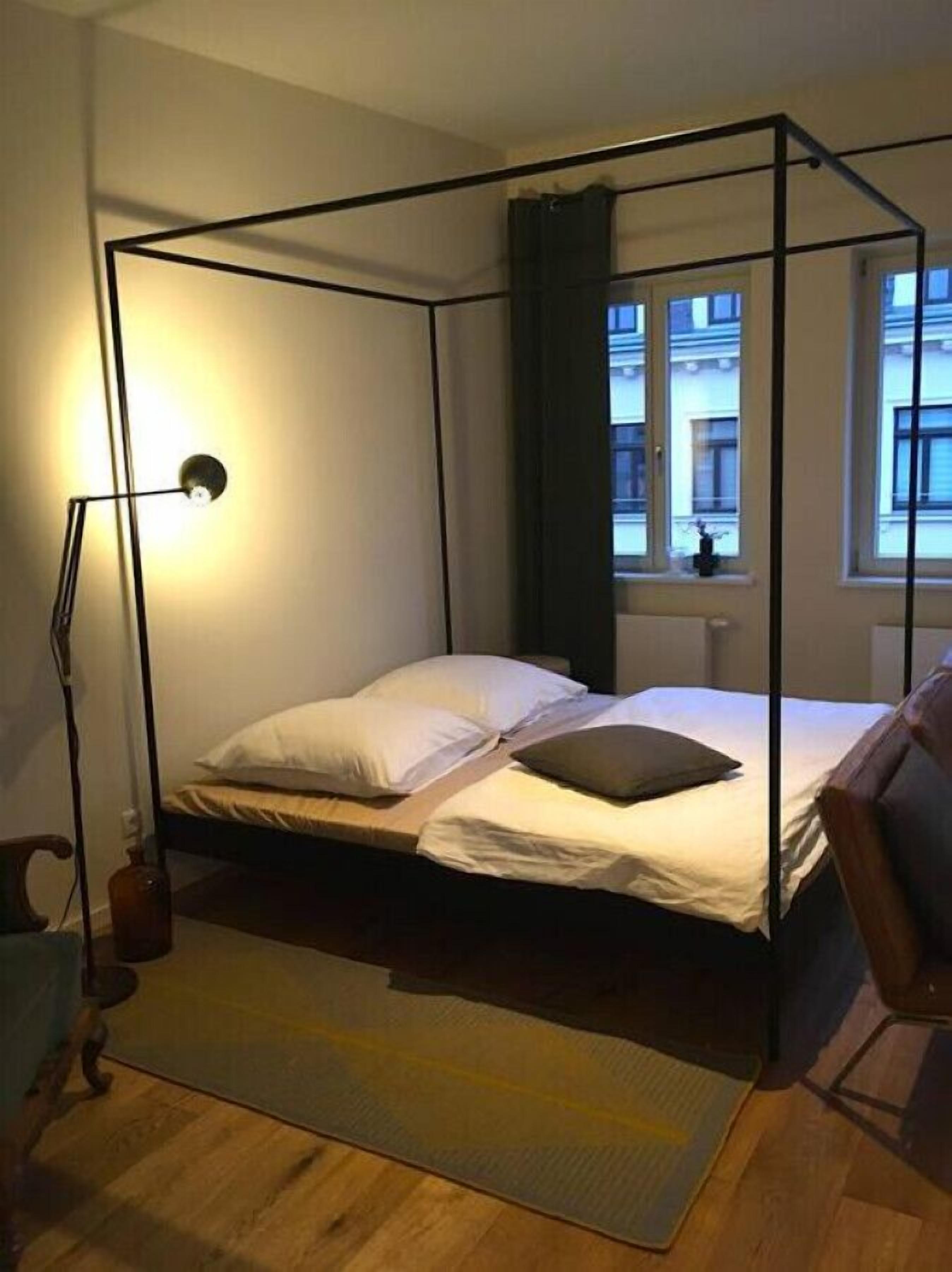 Ein Bett mit Baldachin, ein Nachttisch, eine Lampe und Blick auf Gebäude durch ein Fenster.