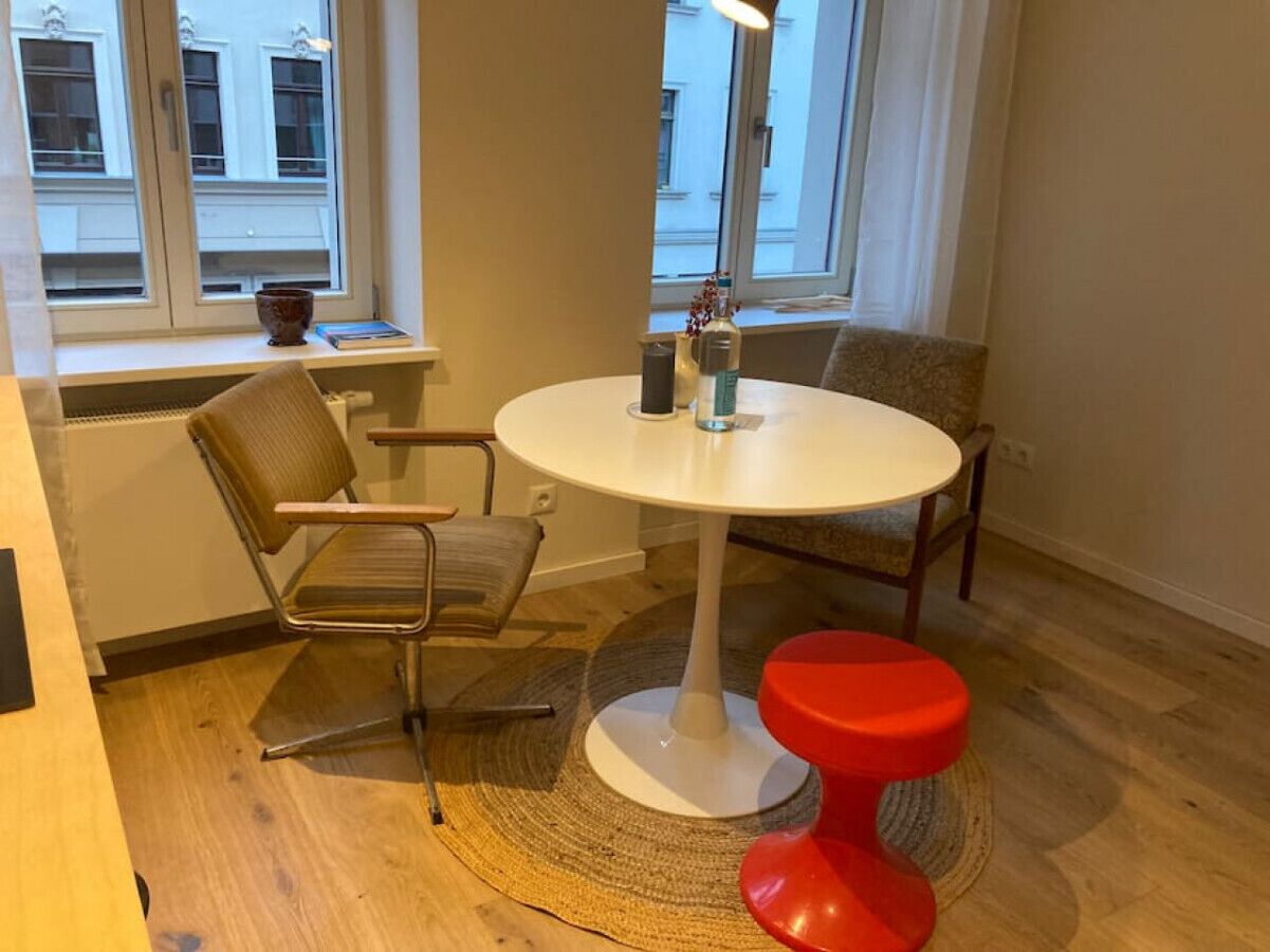 Una stanza con un tavolo rotondo bianco, due sedie, uno sgabello rosso, un tappeto fatto a mano e una finestra che si affaccia su edifici.