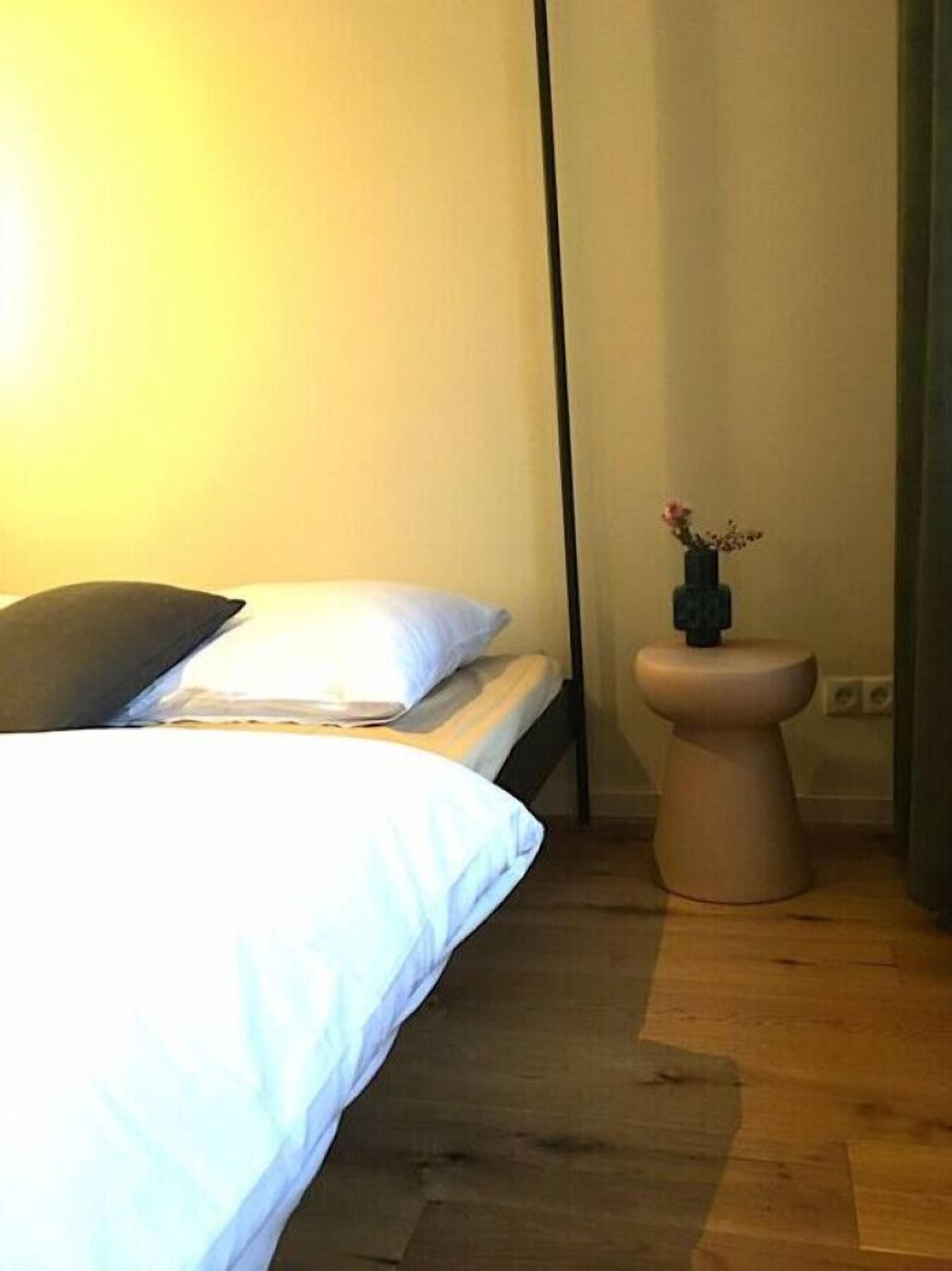 A bedroom with a bed, a bedside table with a vase of flowers, and a wooden floor.