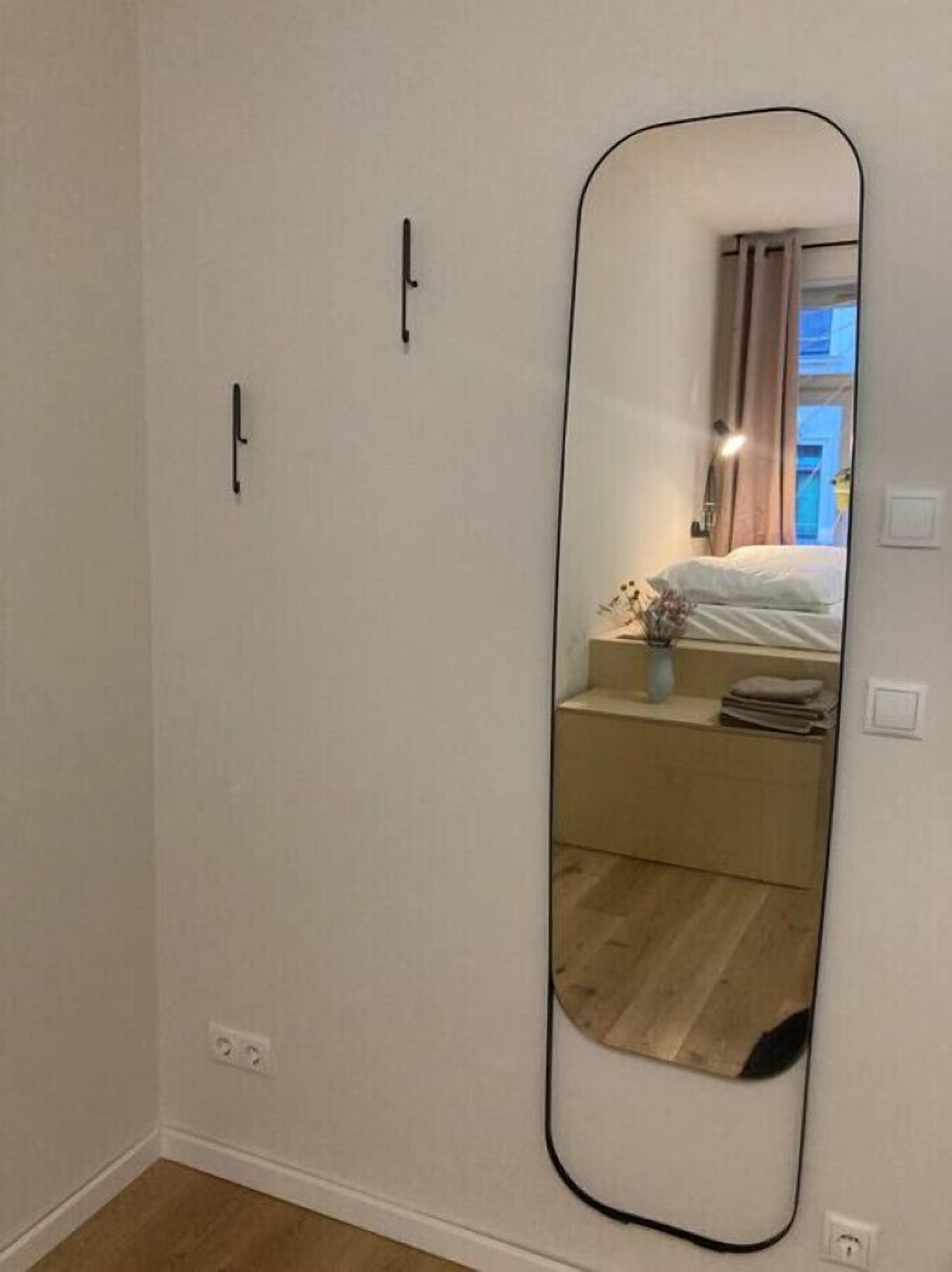 A room with a large mirror, a bed, a nightstand, and a window with curtains.