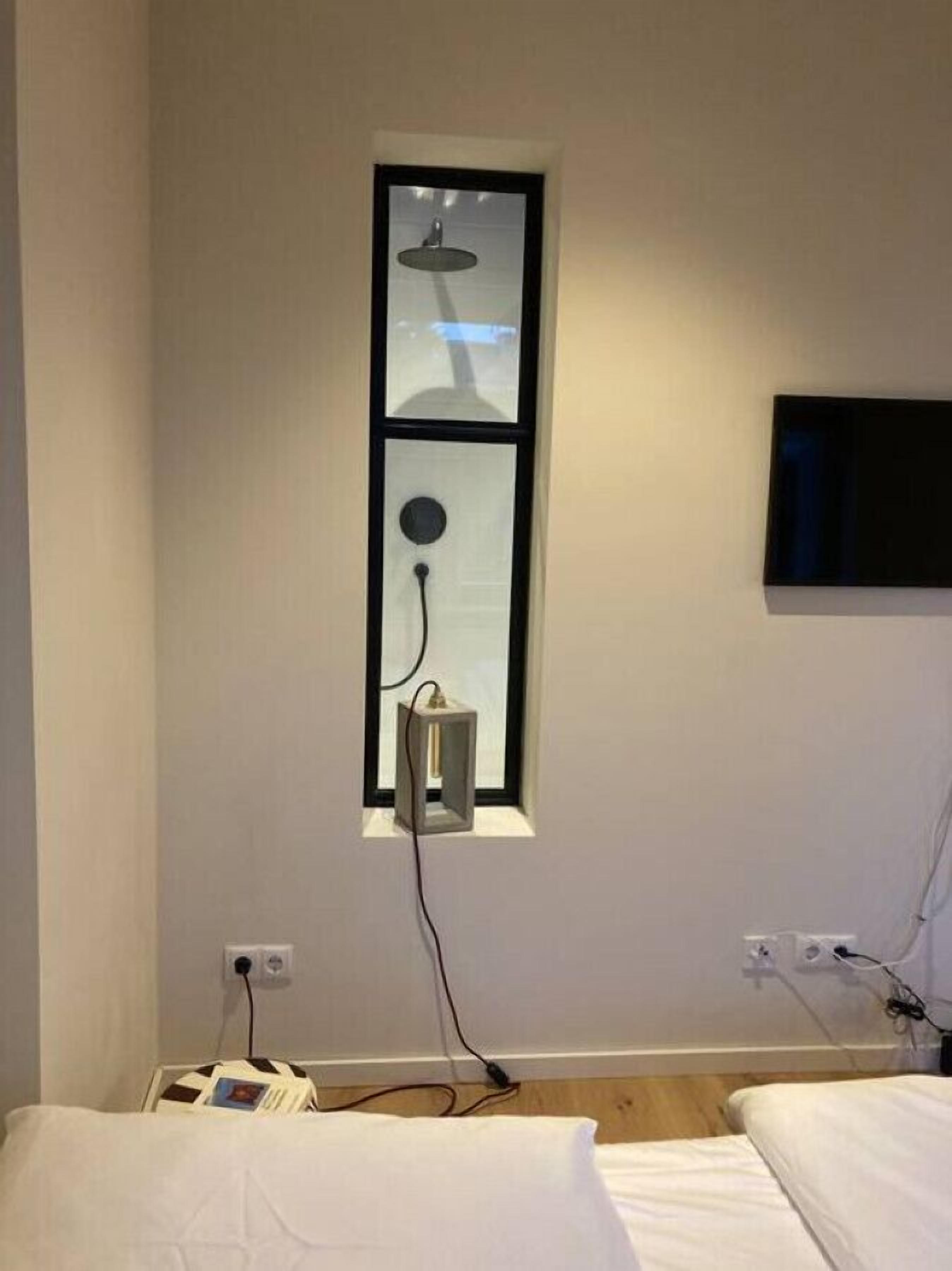 A bedroom with a single bed, a bedside table, a wall-mounted TV, and a window with a light fixture.