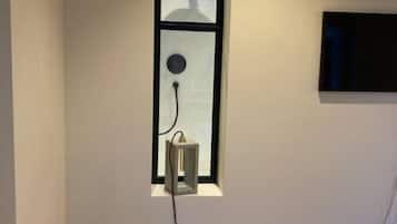 Un dormitorio con una cama individual, una mesita de noche, un televisor montado en la pared y una ventana con lámpara.