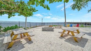 Een terras buiten met picknicktafels en banken, omringd door een hek en bomen, met uitzicht op een waterpartij.