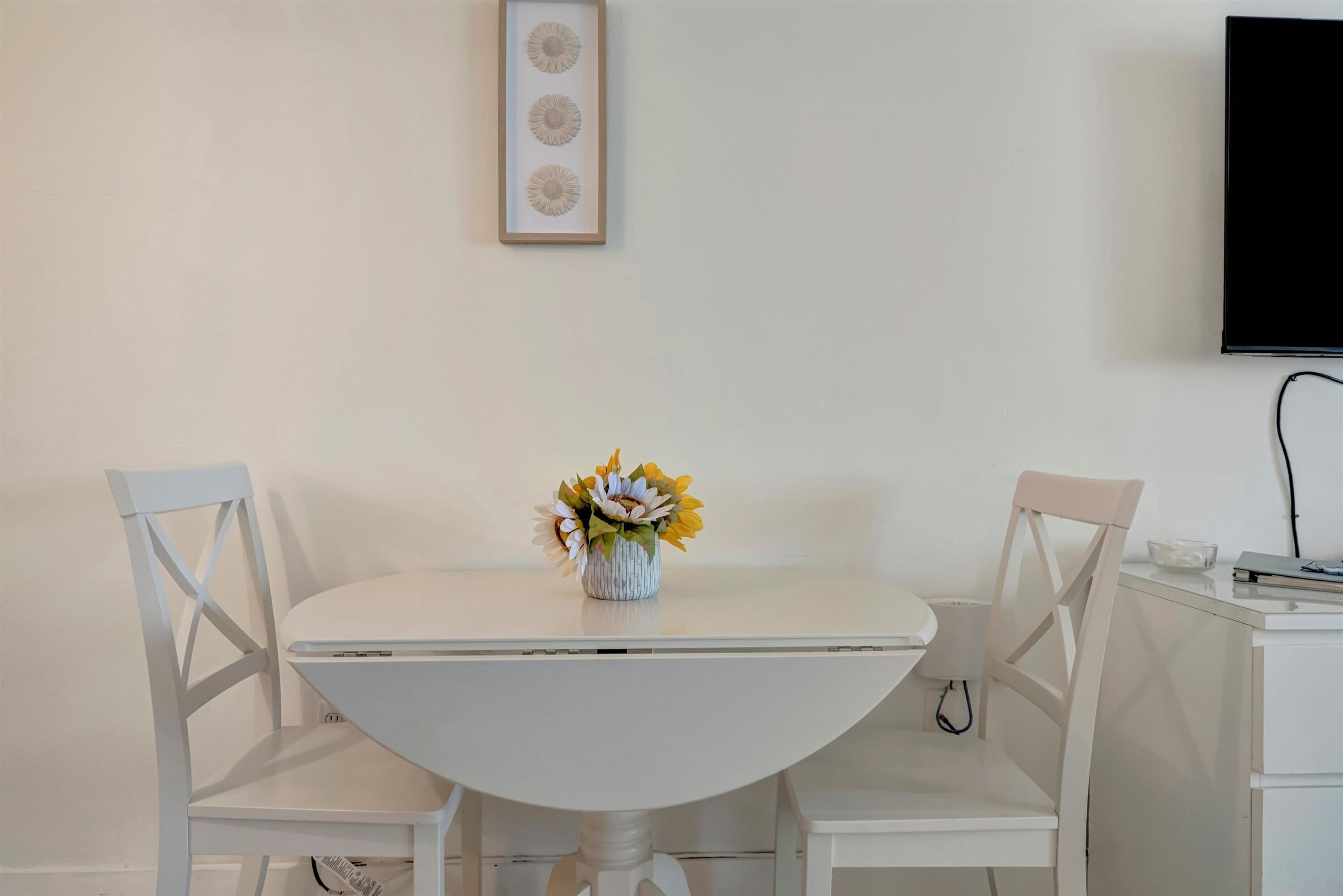 A white dining table with two chairs, a vase with flowers, a framed picture on the wall, and a TV on the wall.