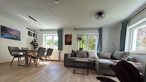 A modern living room with a sectional sofa, a dining table with chairs, and a large window with curtains. - Vacation apartment in a quiet location, directly on the premium hiking trails (Dahn)