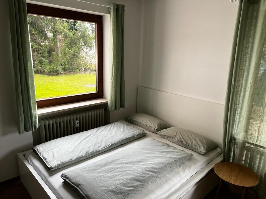 A room with a bed, a window with curtains, a radiator, and a chair.