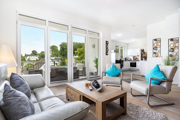 A modern living room with a sofa, armchair, and coffee table, a kitchen in the background, and large sliding glass doors leading to an outdoor area.