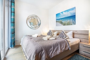 A bedroom with a bed, a wooden headboard, a wall-mounted mirror, a painting of a beach, and a window with curtains.