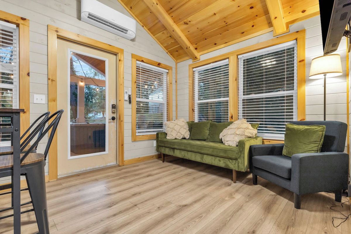 A cozy interior with a wooden ceiling, a green sofa, a grey armchair, and a dining area with a chair and table.