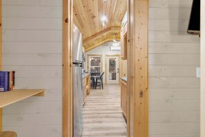 A narrow hallway with wooden walls and a sloped ceiling, leading to a dining area with a table and chairs.