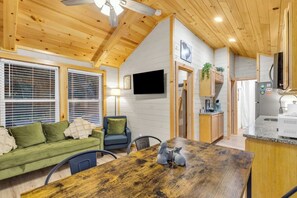 A cozy cabin interior with a wooden ceiling, a dining table, a sofa, a TV mounted on the wall, and a kitchen area.
