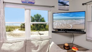 A room with a large window overlooking a beach, a flat-screen TV mounted on the wall, a wooden table with a coconut and a fish decoration, and a couch with a 'Welcome to Paradise' sign above it. - Beachside at Birdsong #17 Pool 5 Steps to the Sand (Treasure Island)