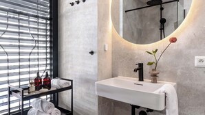 A modern bathroom with a white sink, black faucet, and a round mirror with a light. There is a towel rack with towels and a shelf with toiletries. - TurnKey | Henry's Tower Apartment II-3 (Hlavní město Praha)