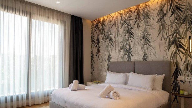 Una habitación de hotel con una cama grande, una mesita de noche, una lámpara y un ventanal con cortinas translúcidas.