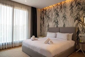 A hotel room with a large bed, a nightstand, a lamp, and a window with sheer curtains. - Kennah Catalina | Silver Palm, New Cairo - Families Only (New Cairo)