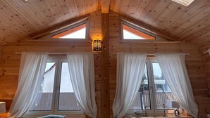 Eine Holzhütte mit zwei Dachfenstern, großen Fenstern mit weißen Vorhängen und einem kleinen Tisch mit Lampe und Dekoration.