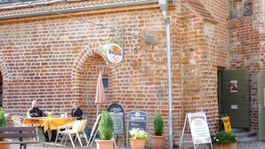 Ein Backsteinhaus mit einem Schild, auf dem 'Burgcafé in Spree' steht. Es gibt Tische und Stühle im Freien, Topfpflanzen und ein Schild für das Oktoberfest.