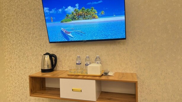 Ein an der Wand befestigter Fernseher zeigt eine tropische Inselansicht, ein Holzboden mit Wasserkocher, Gläsern, Flaschen und einer kleinen weißen Schachtel.