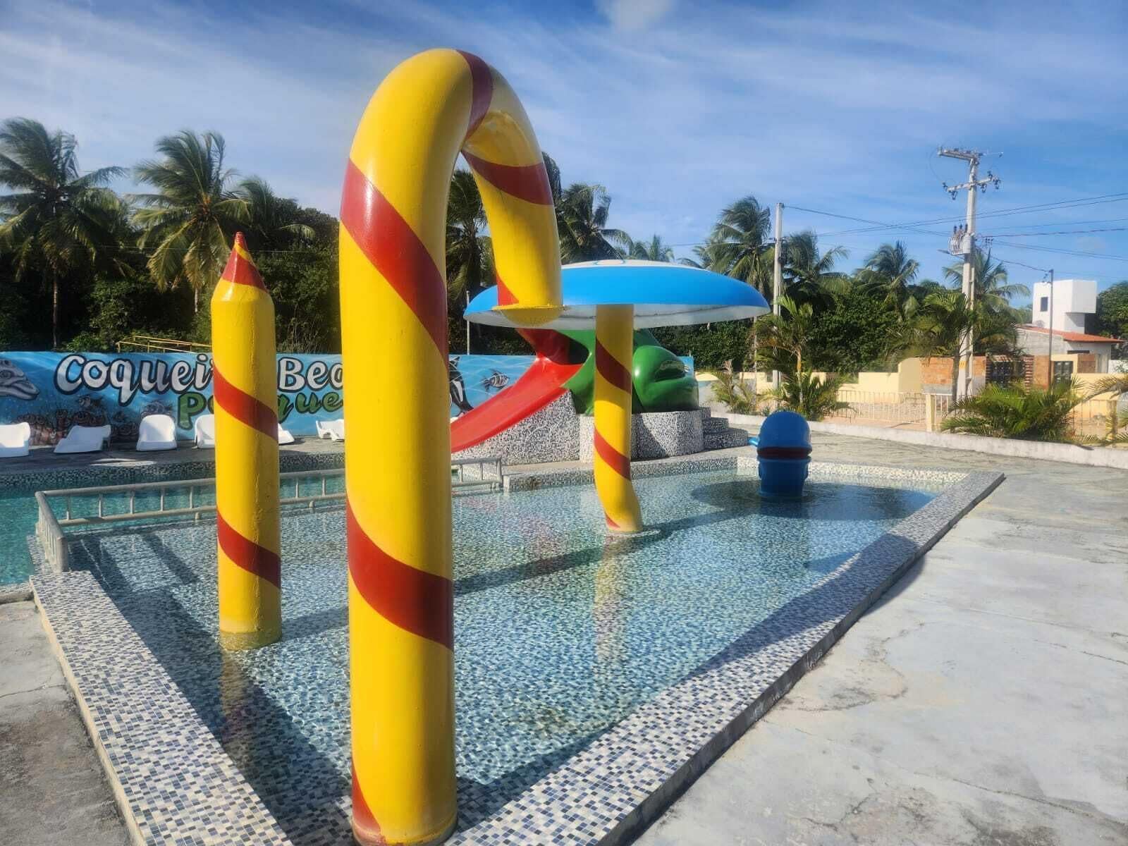A swimming pool area with a water slide, a lounge area with white chairs, and a sign that reads "Coquei Beach".