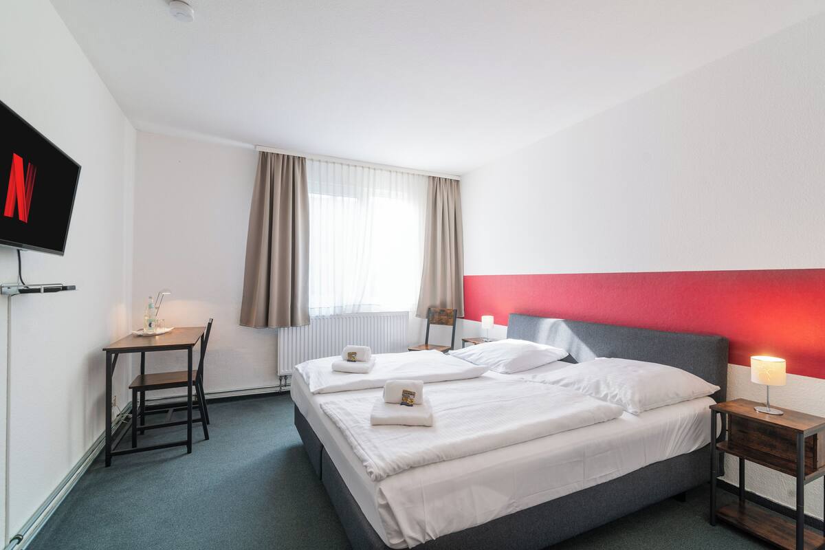 Ein Hotelzimmer mit zwei Betten, einem Schreibtisch, einem Fernseher und einem Fenster mit Vorhängen.