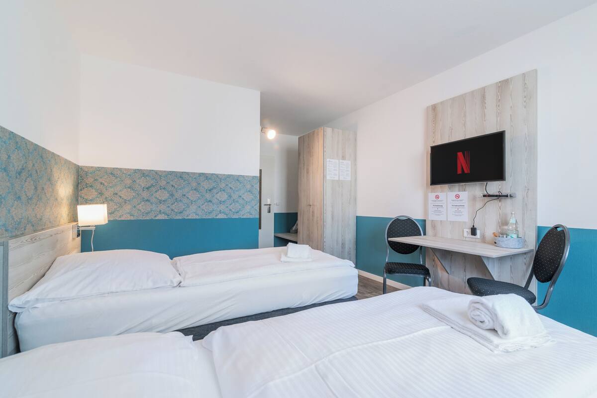 Ein Hotelzimmer mit zwei Betten, einem Schreibtisch, einem Sessel, einem Fernseher und einem Wandregal.