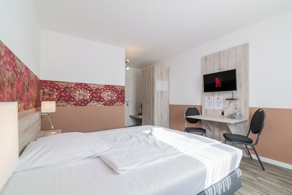 A hotel room with a bed, a desk, a chair, a TV, and a wall-mounted shelf.