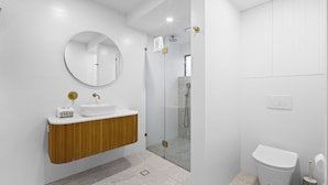 A modern bathroom with a wooden vanity, a round mirror, a toilet, and a glass-enclosed shower area. - Beachside Bliss on Palm Avenue (Surfers Paradise)