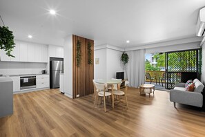 A modern kitchen with white cabinets, a wooden accent wall, and a dining area with a round table and chairs. - Beachside Bliss on Palm Avenue (Surfers Paradise)