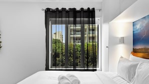 A hotel room with a bed, a window with black curtains, a view of greenery and buildings, and a wall-mounted lamp. - Beachside Bliss on Palm Avenue (Surfers Paradise)