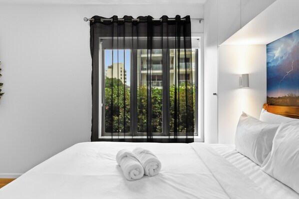 A hotel room with a bed, a window with black curtains, a view of greenery and buildings, and a wall-mounted lamp.
